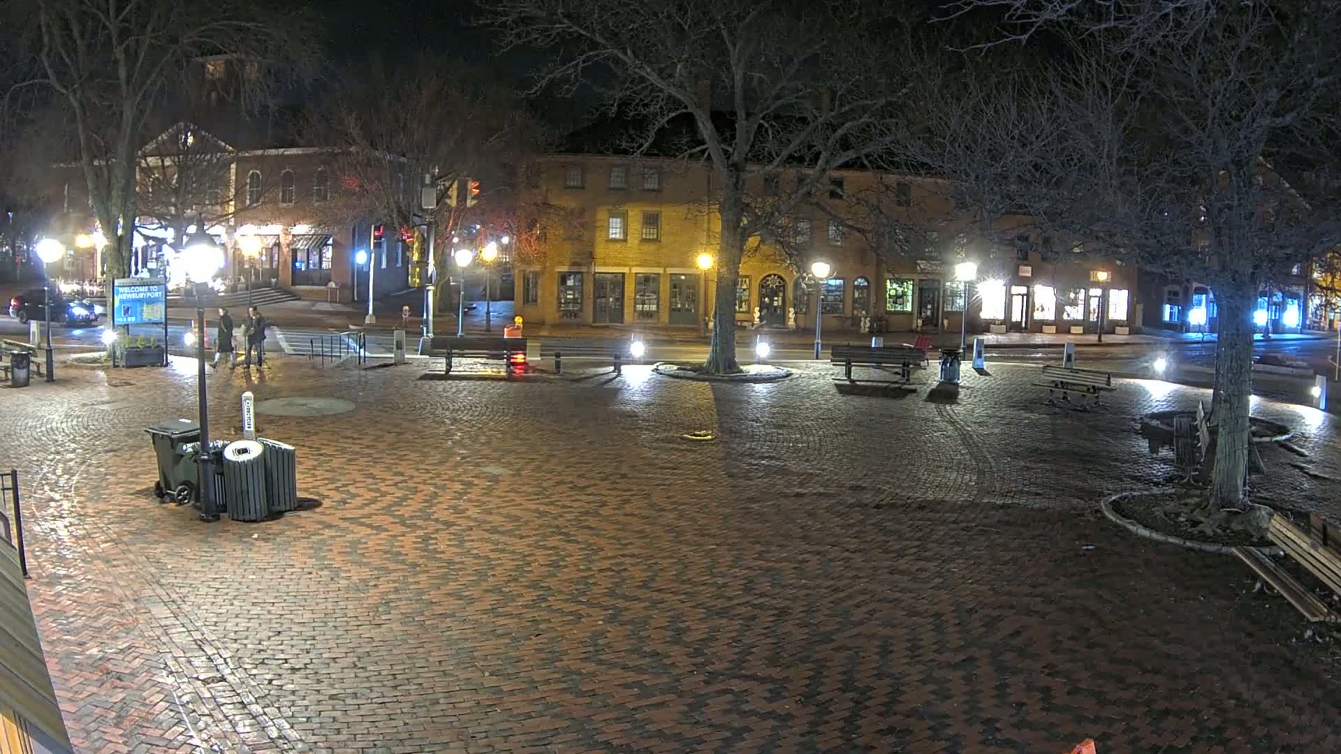 A brick town square at night, illuminated by streetlights, with a few people walking and empty park benches around a central tree.