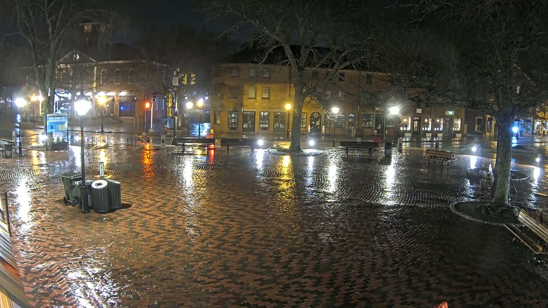 A rain-slicked brick town square at night is illuminated by streetlights reflecting on the wet cobblestones, with buildings and trees lining the perimeter.