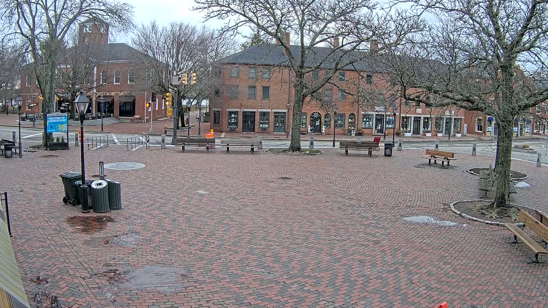 A brick plaza with several benches sits in front of brick buildings under a cloudy sky.