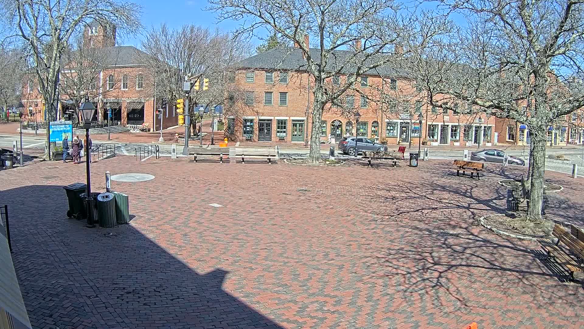 A brick town square with benches and a few people, under a sunny, clear sky, is surrounded by brick buildings with shops on the ground floor.