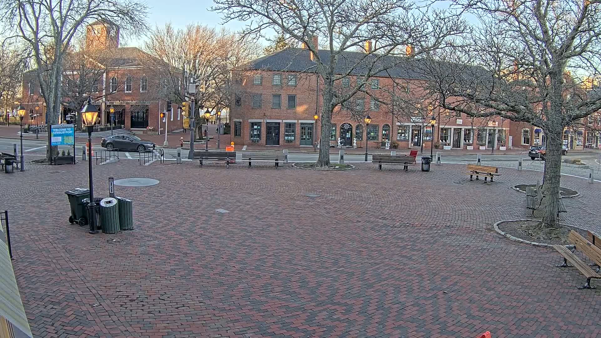 A brick plaza with several benches and trees, situated in front of brick buildings on a clear, late afternoon.