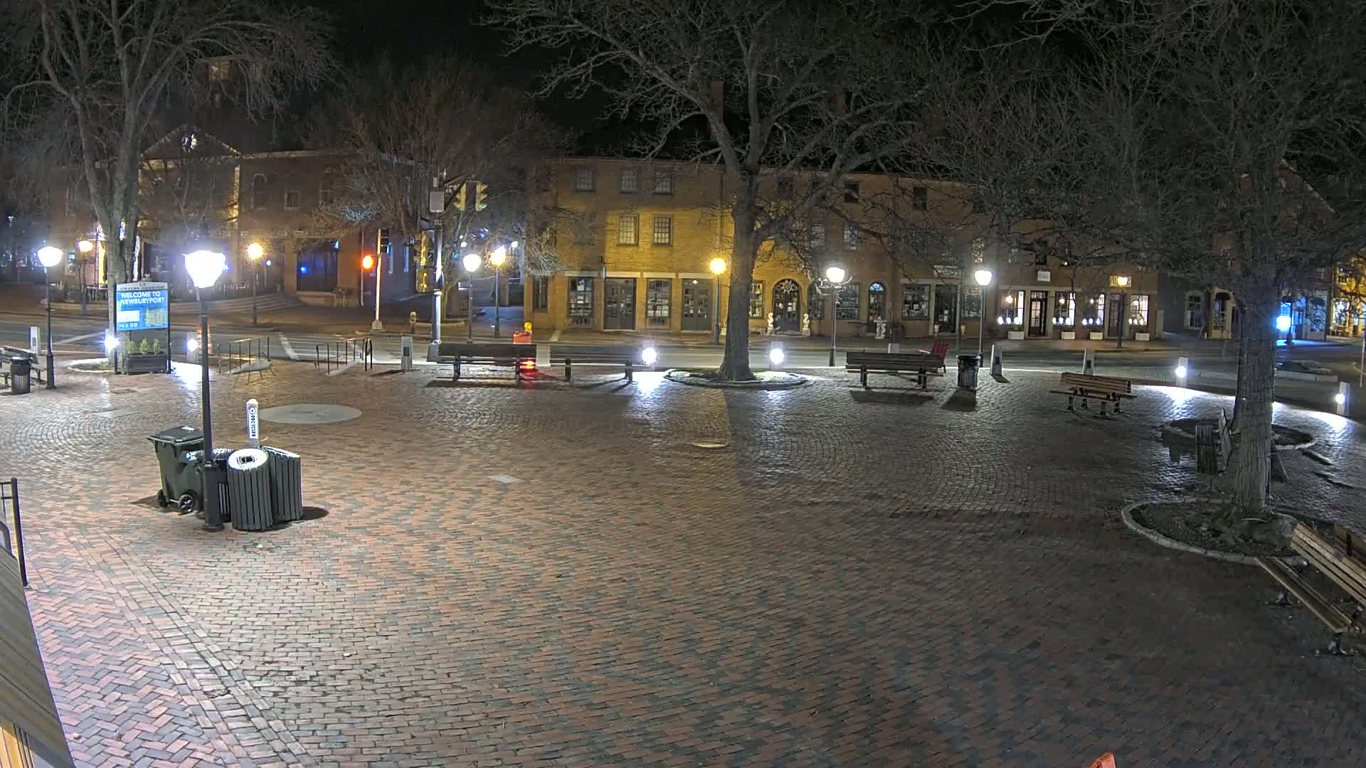 A brick town square at night, illuminated by streetlights, with benches, trees, and buildings lining the perimeter.