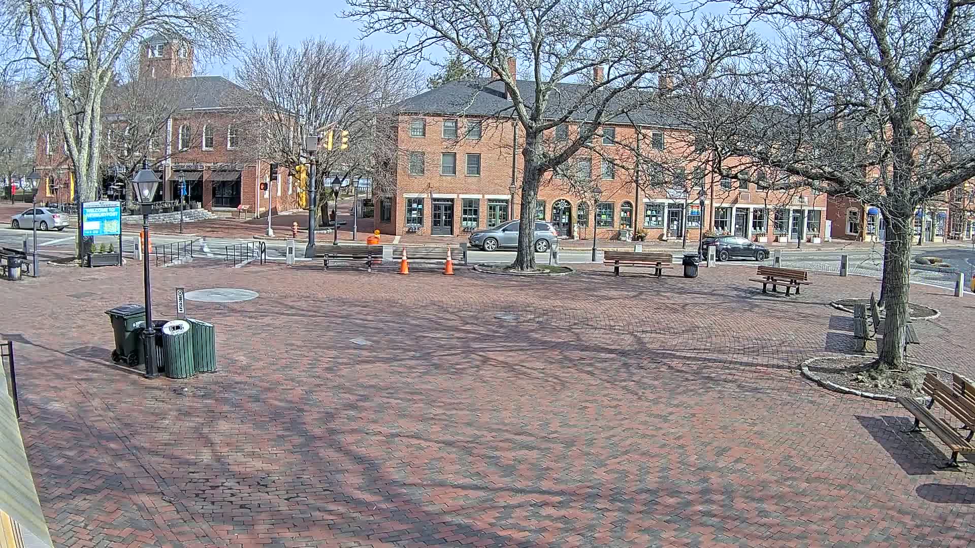 A brick town square with benches and bare trees under a sunny sky, features several brick buildings across a street.