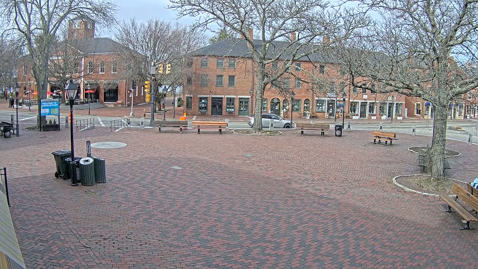 A brick plaza with benches and trash cans is surrounded by brick buildings and bare trees under a cloudy sky.