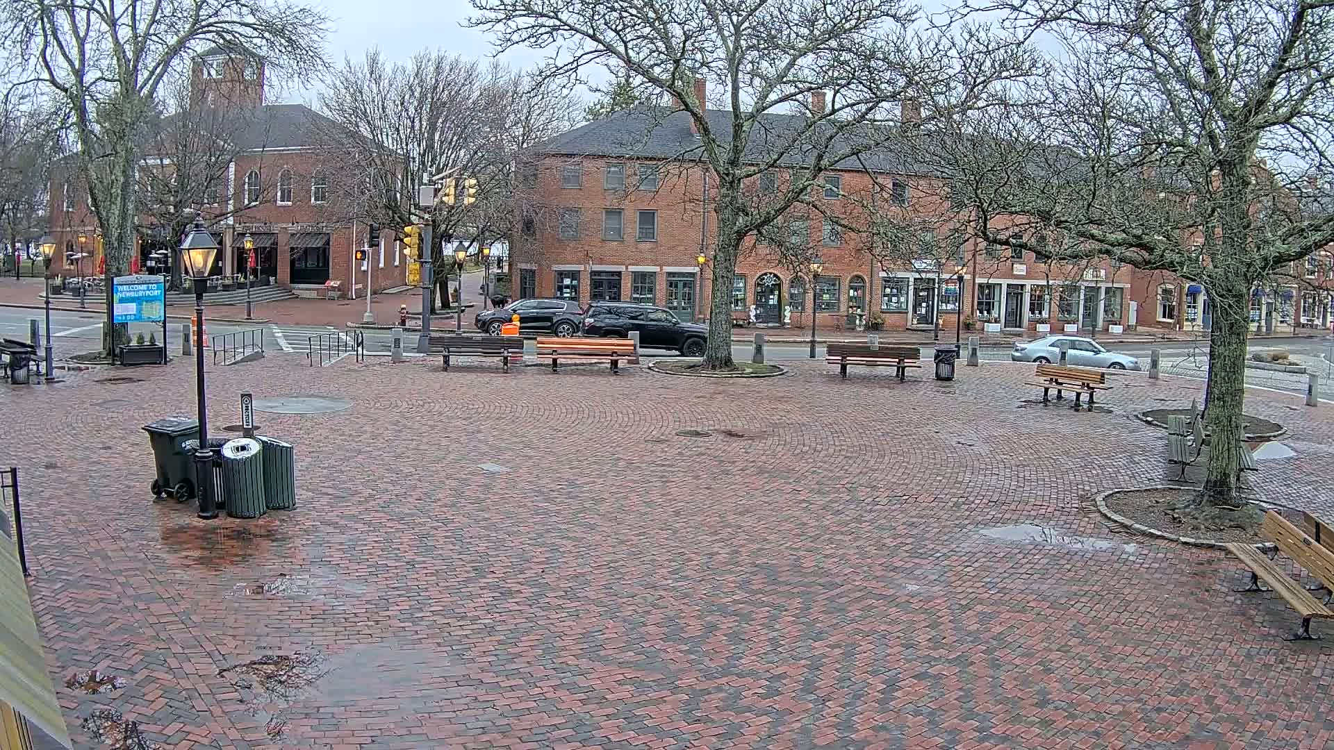 A brick town square with benches and several brick buildings is wet from recent rain under a cloudy sky.