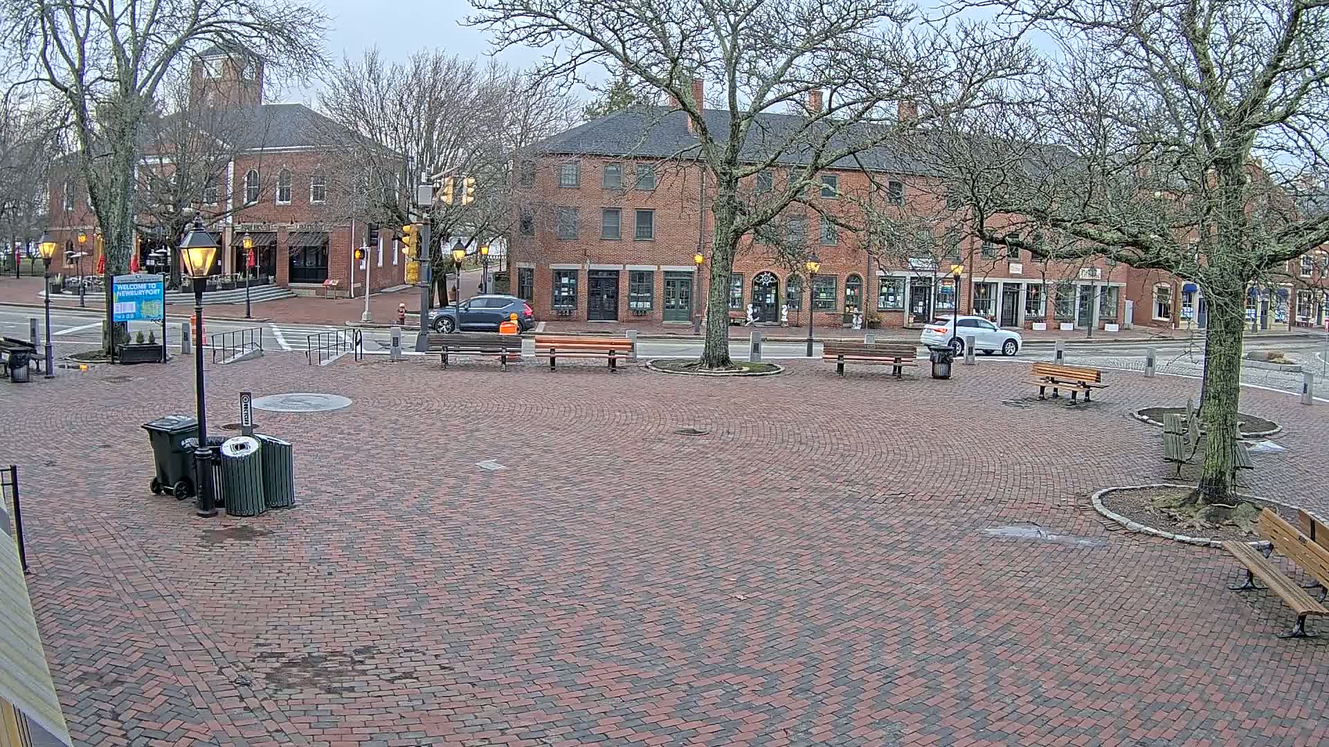 A brick town square with several benches and trash cans, under an overcast sky, is surrounded by brick buildings and bare trees.