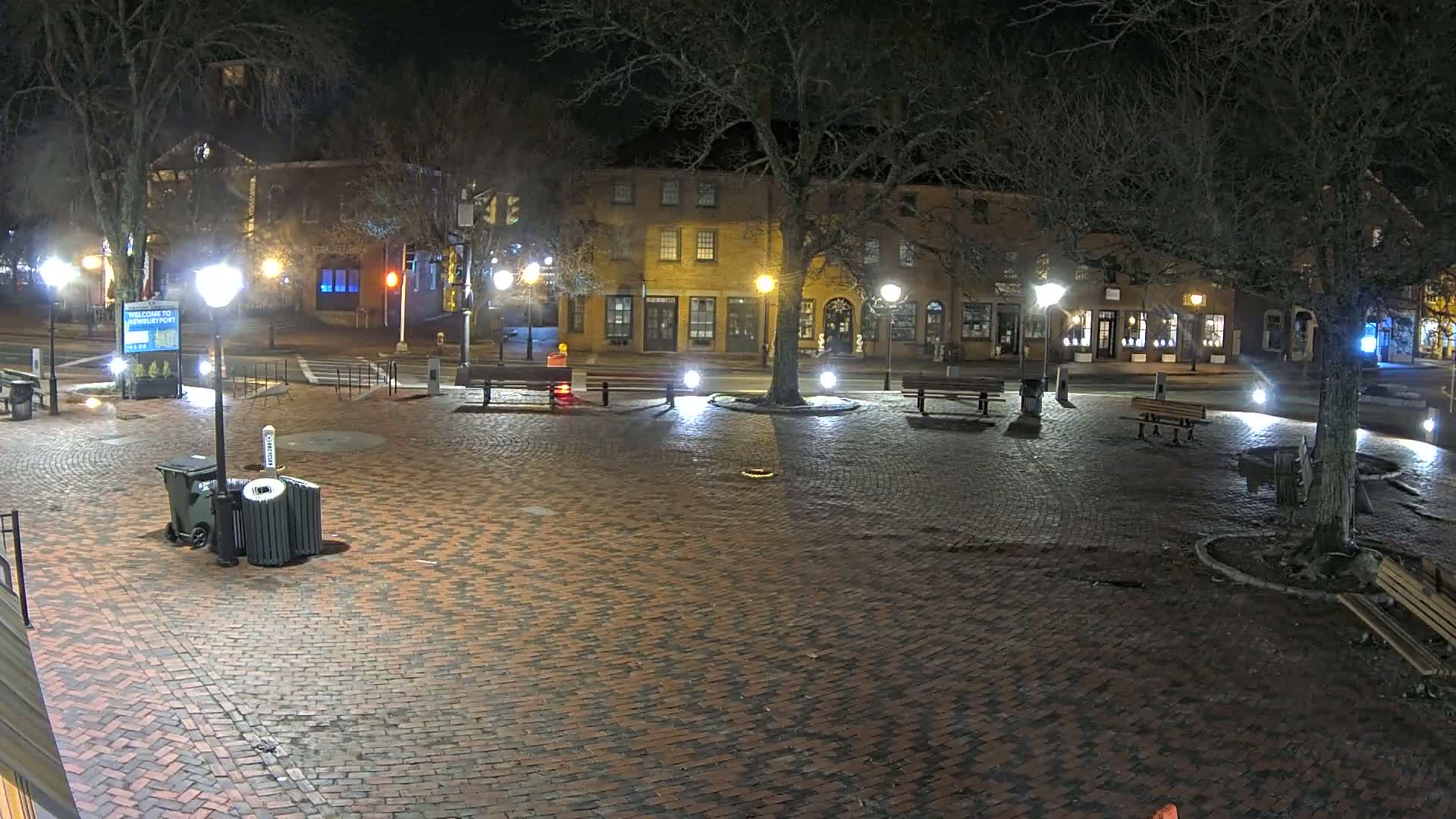 A brick town square at night, illuminated by streetlights, is surrounded by several low buildings and bare trees.