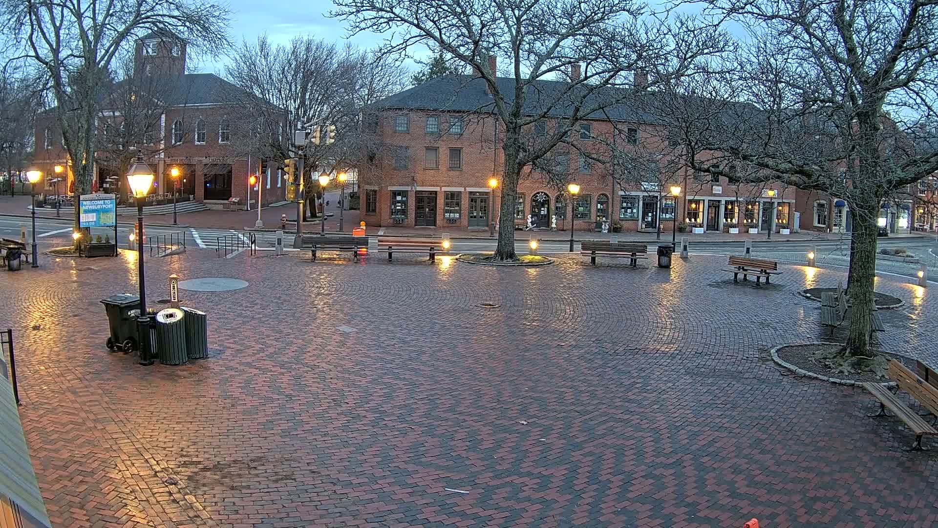 A brick town square at dusk, illuminated by lampposts, with benches and trees surrounding a central, wet paved area.