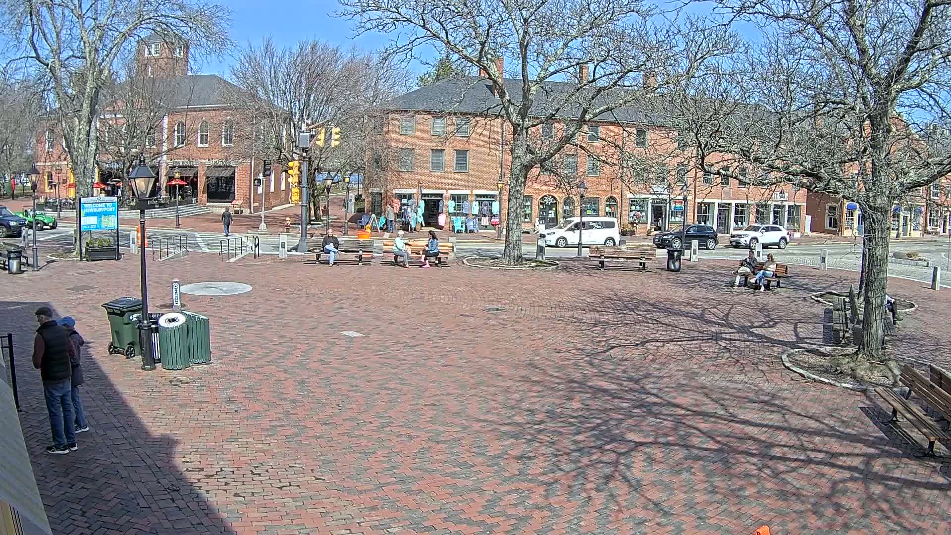 A brick town square with several people sitting on benches under leafless trees on a sunny day.