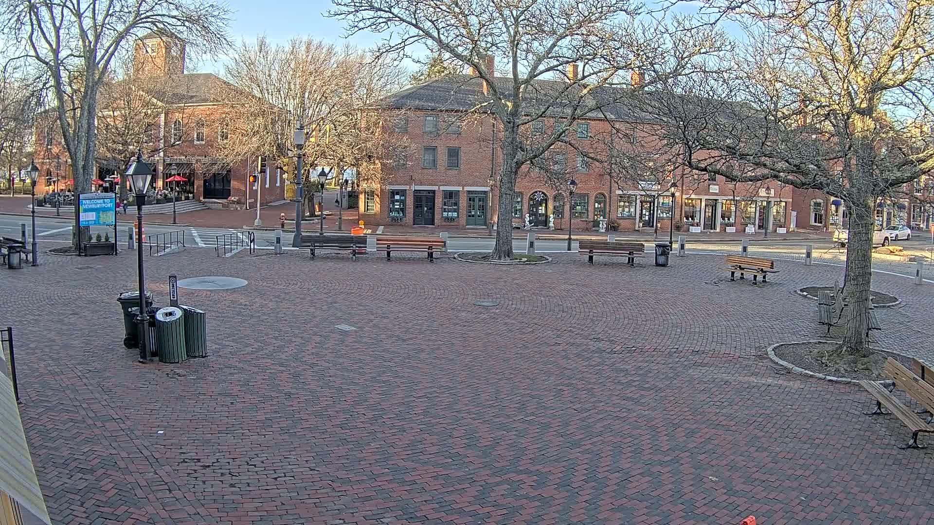 A brick town square with several benches and trees, under a sunny, clear sky, is surrounded by brick buildings with storefronts.