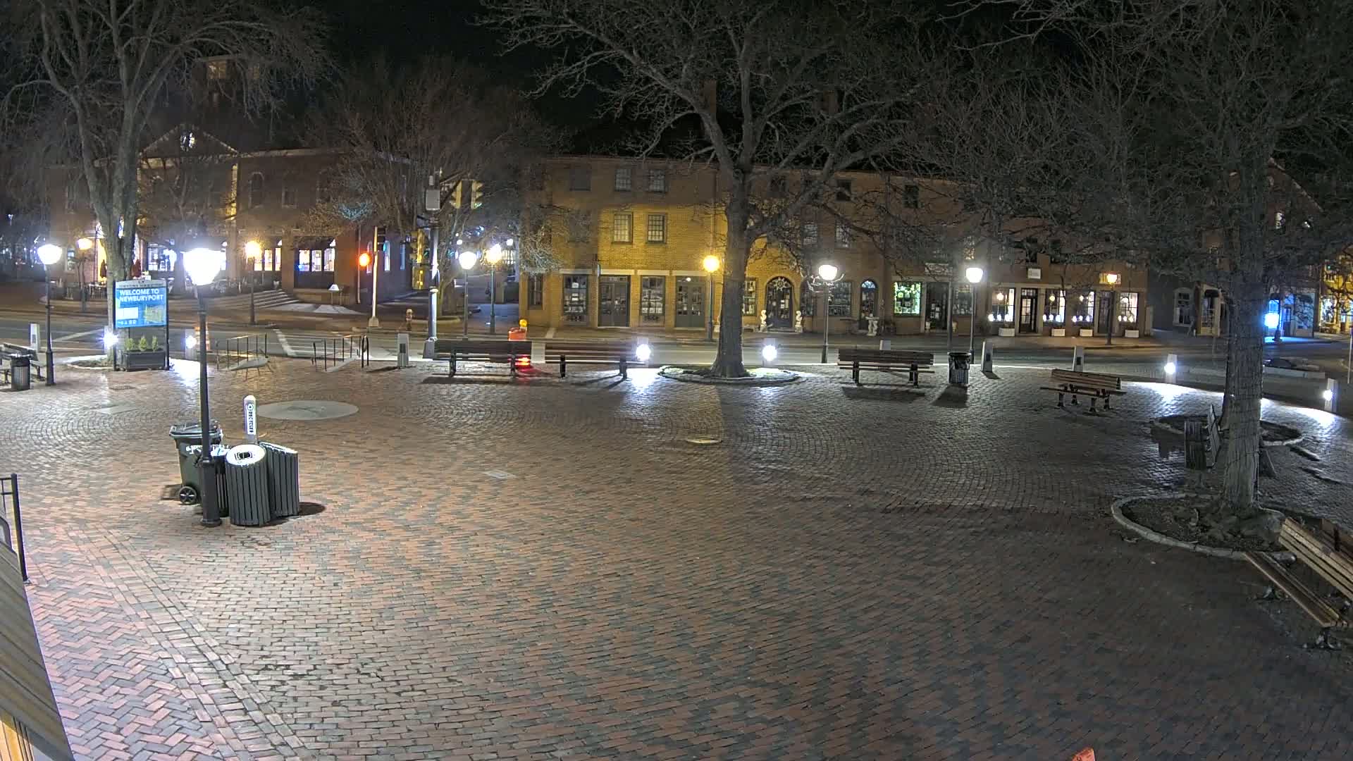 A brick town square at night, illuminated by streetlights, features benches, trees, and a row of buildings along the perimeter.