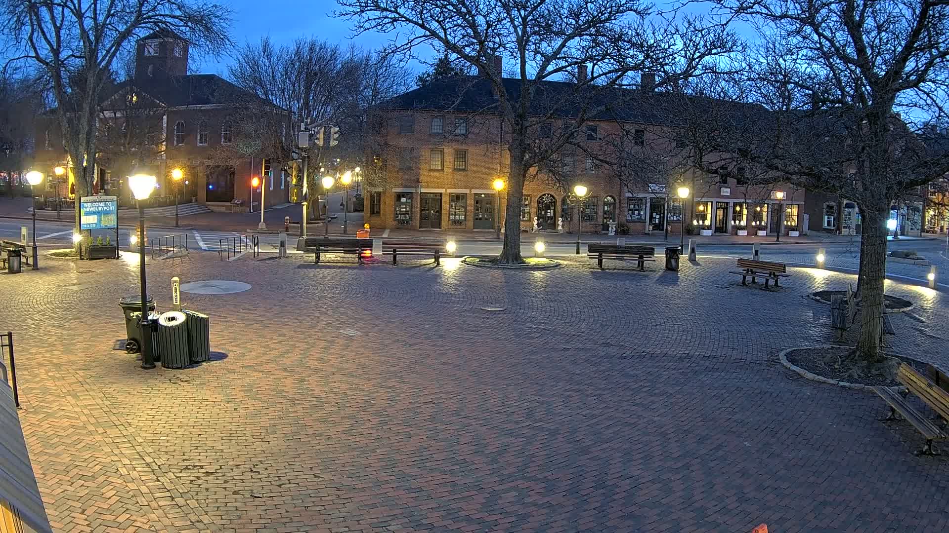 A brick town square at dusk, illuminated by lampposts, features several benches and trees surrounding a brick-paved area in front of brick buildings.