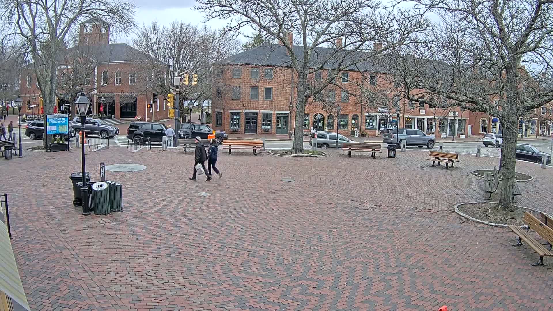 A brick plaza with benches and several parked cars is surrounded by brick buildings under an overcast sky.