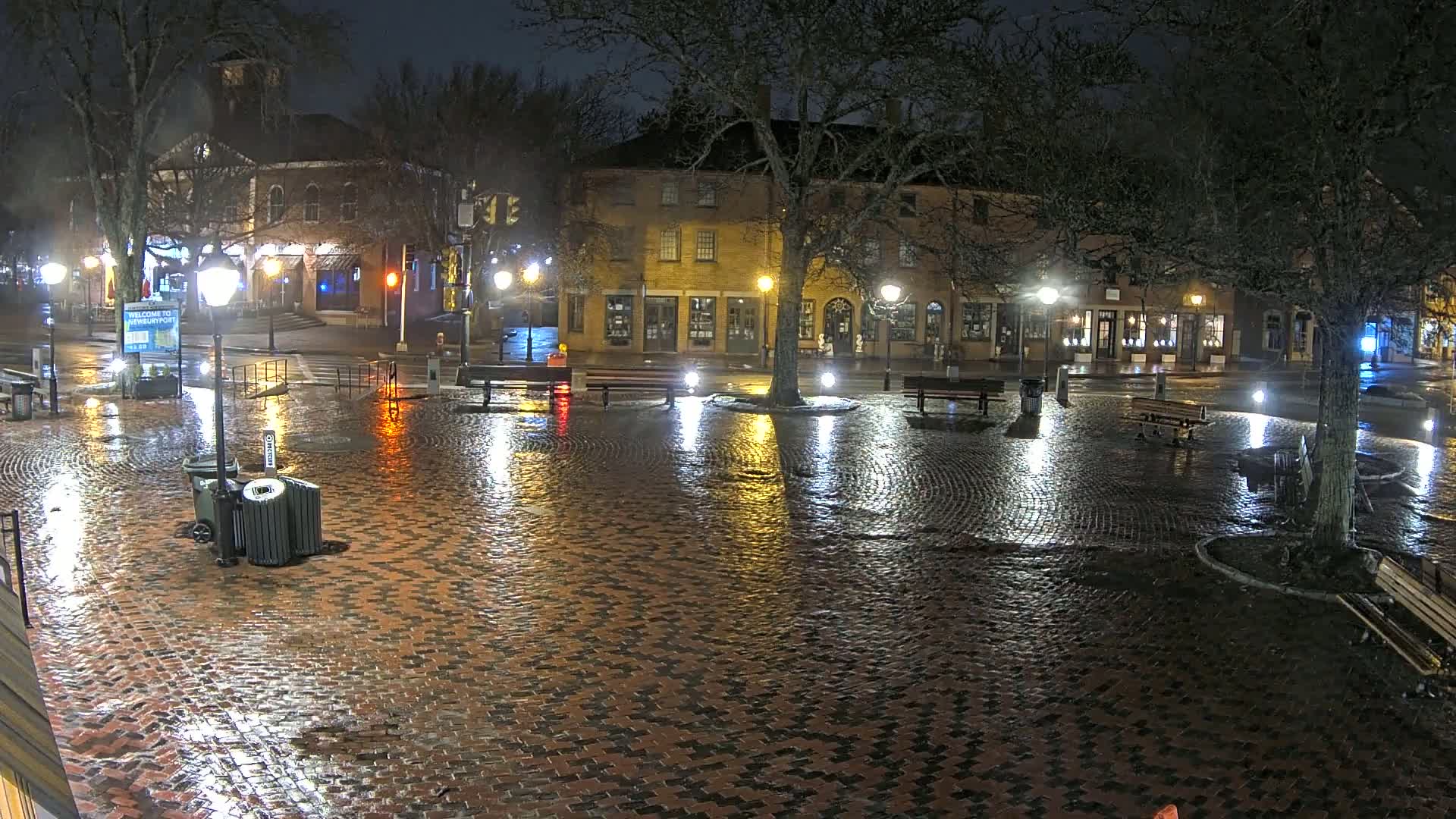A rain-slicked brick town square at night, illuminated by streetlights, features several benches and buildings lining the perimeter.