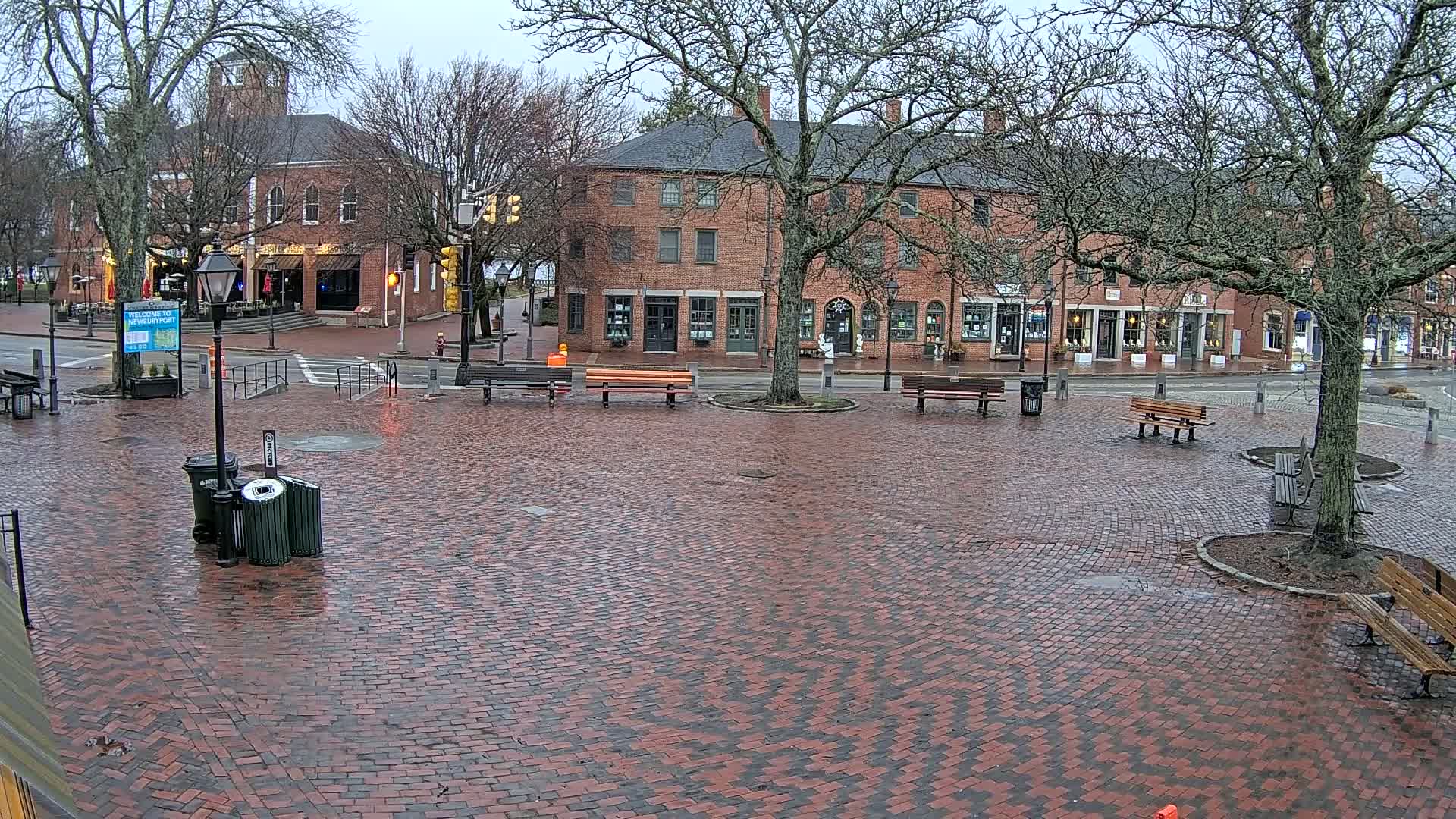 A brick town square with several benches and buildings is shown on a wet, overcast day.