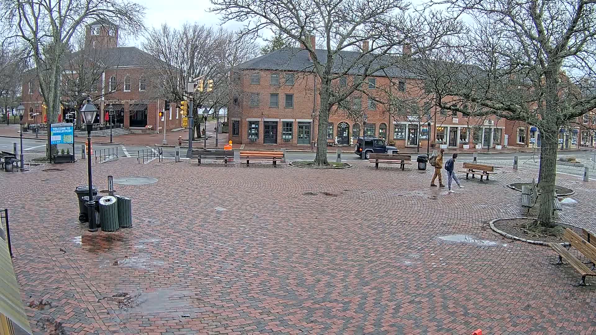 A brick plaza, with benches and trees, sits between two brick buildings under an overcast sky, and two people walk across it.