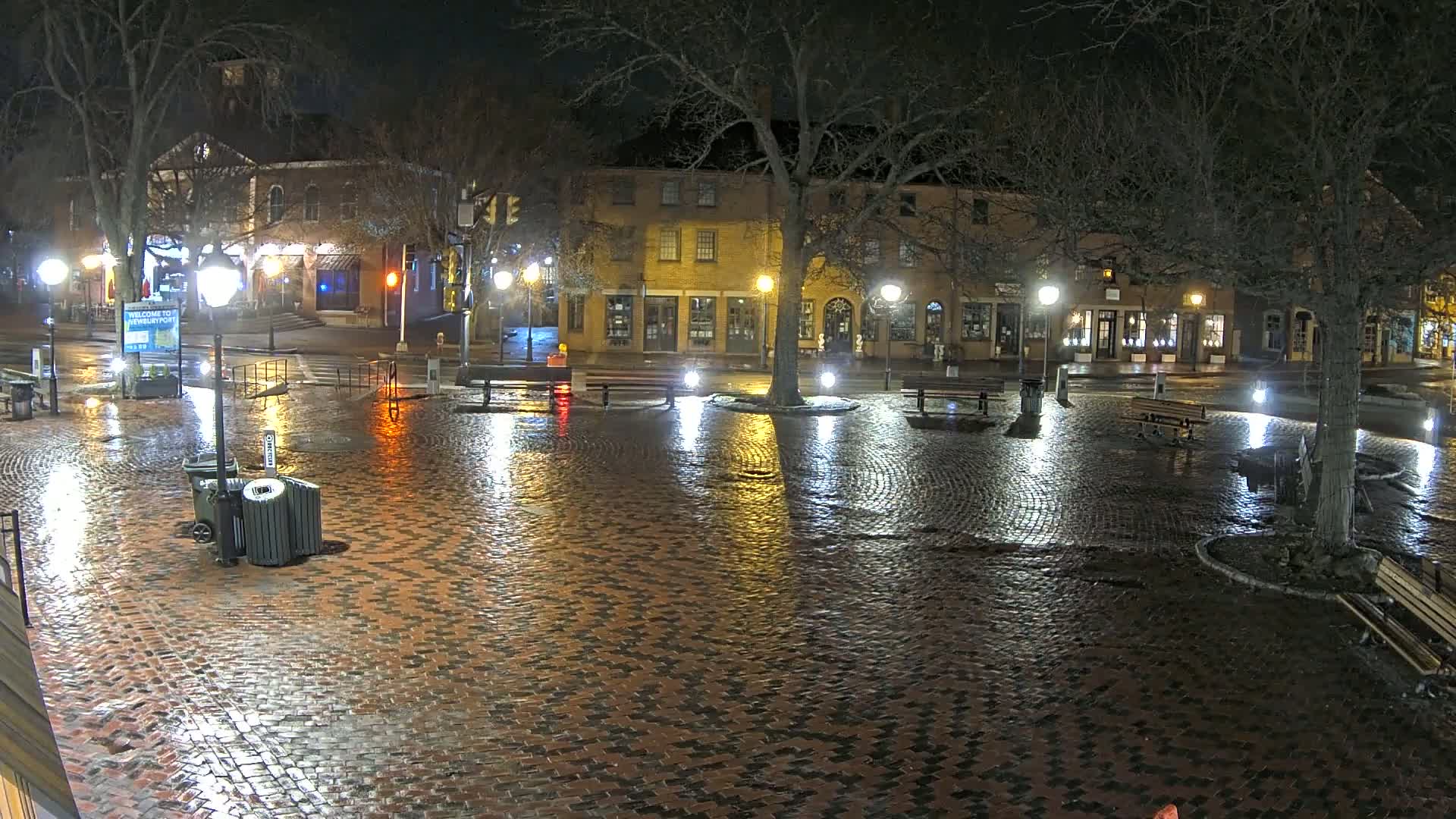 A rain-slicked brick town square at night, illuminated by streetlights, features several benches and trees, with buildings lining the perimeter.