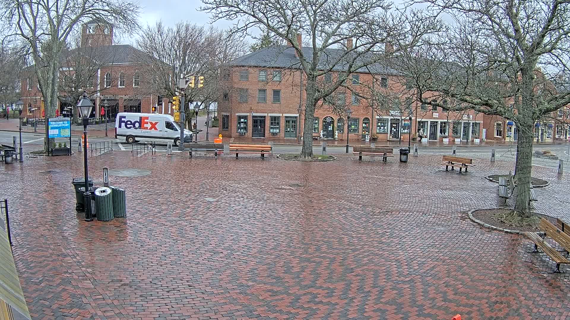 A brick town square with several benches and a FedEx van is seen on a wet, overcast day.