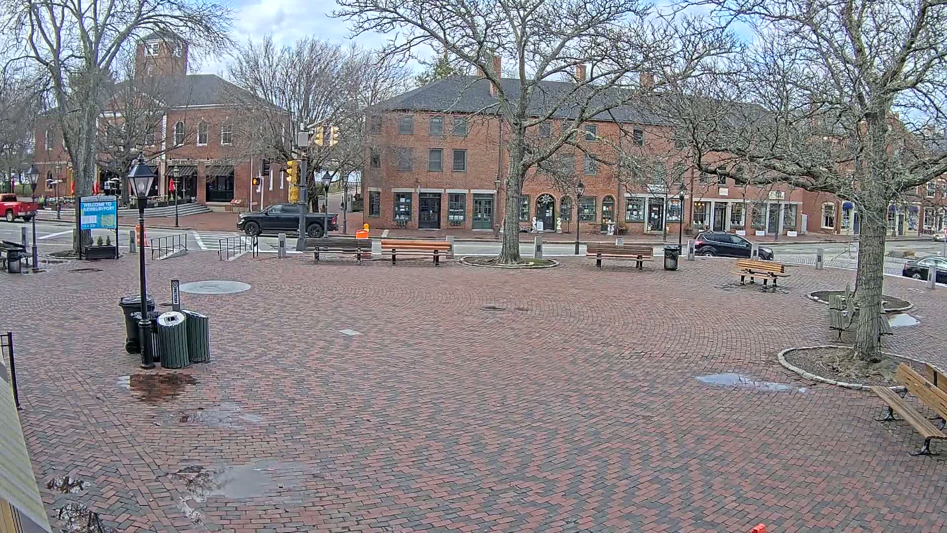 A brick town square with benches and trees under a partly cloudy sky, surrounded by brick buildings.