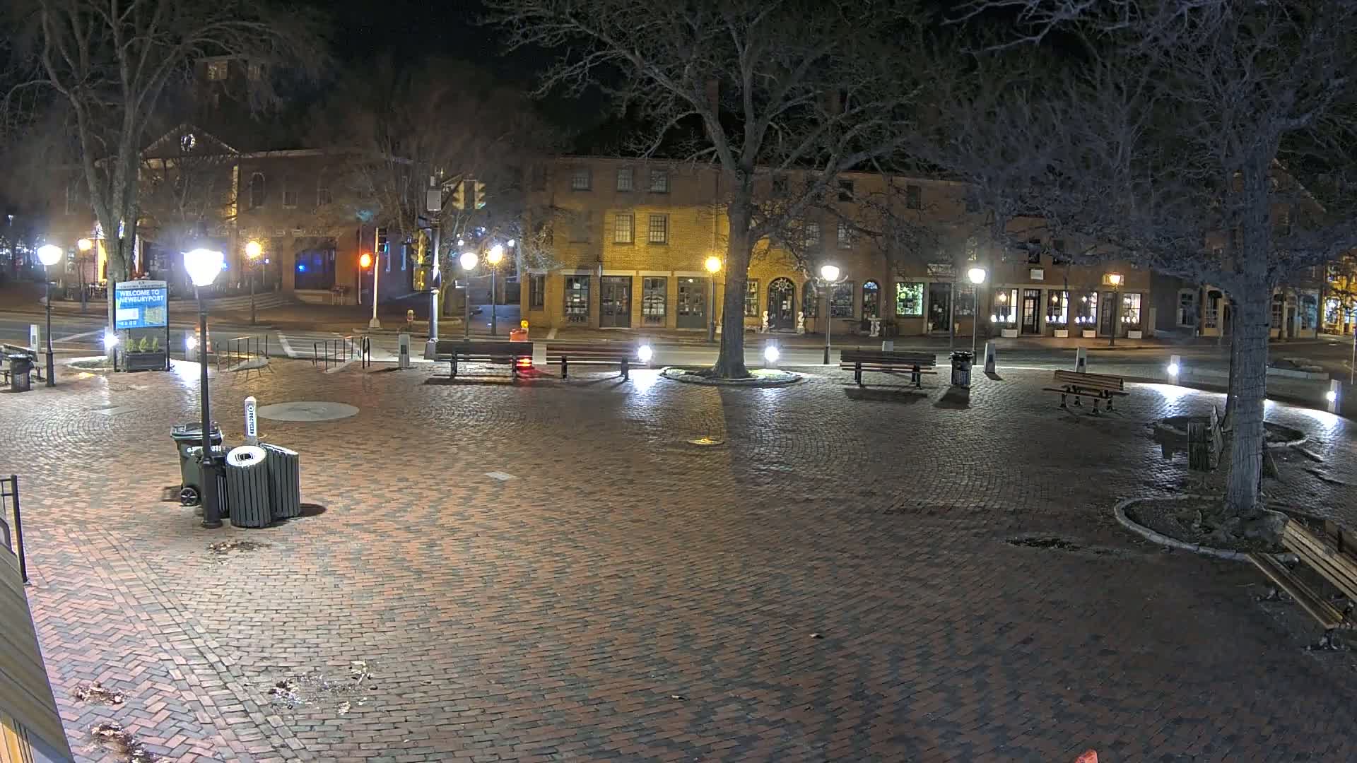 A brick plaza at night, illuminated by streetlights, with benches and a tree in the center, and brick buildings lining the street beyond.