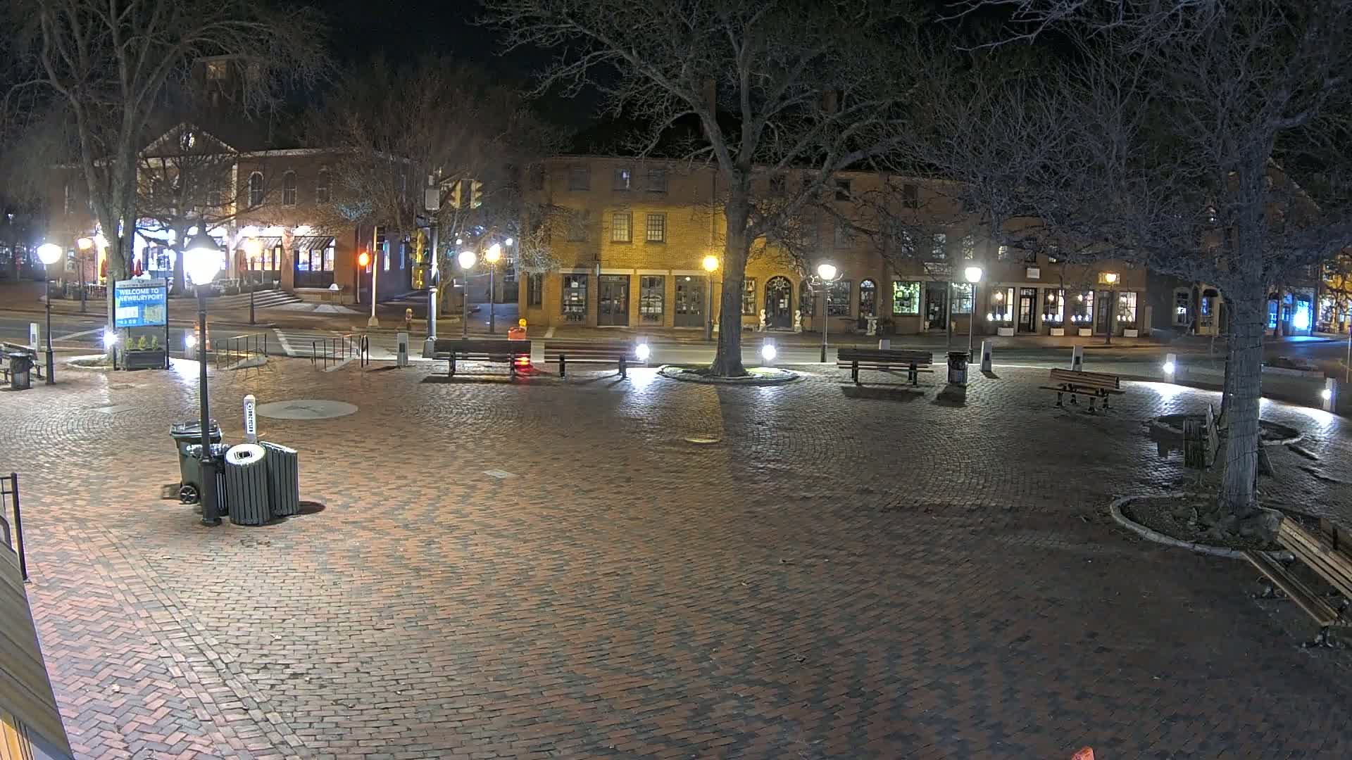 A brick town square at night, illuminated by streetlights, with benches and trees surrounding a central area, and buildings lining the street in the background.
