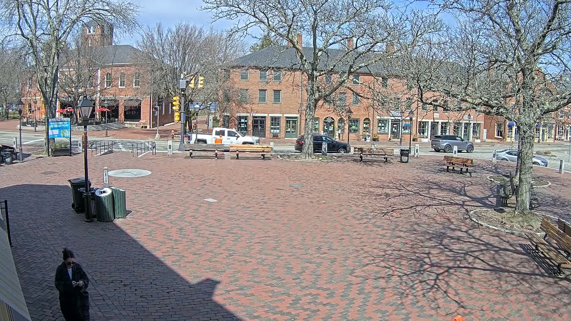 A brick town square with benches, a few cars, and bare trees under a sunny sky.