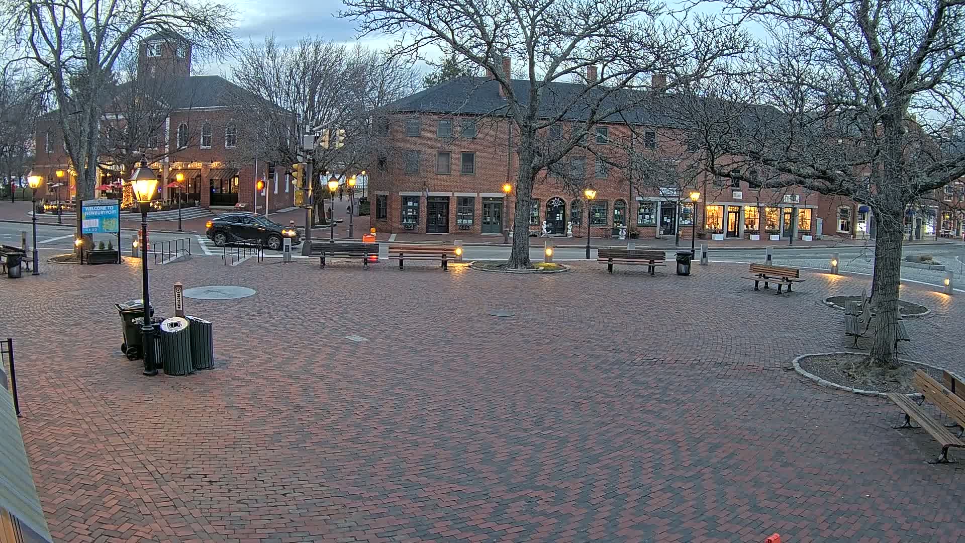A brick town square at dusk, under a mostly clear sky, is surrounded by brick buildings with lit windows and sparsely populated by several park benches and a few pedestrians.