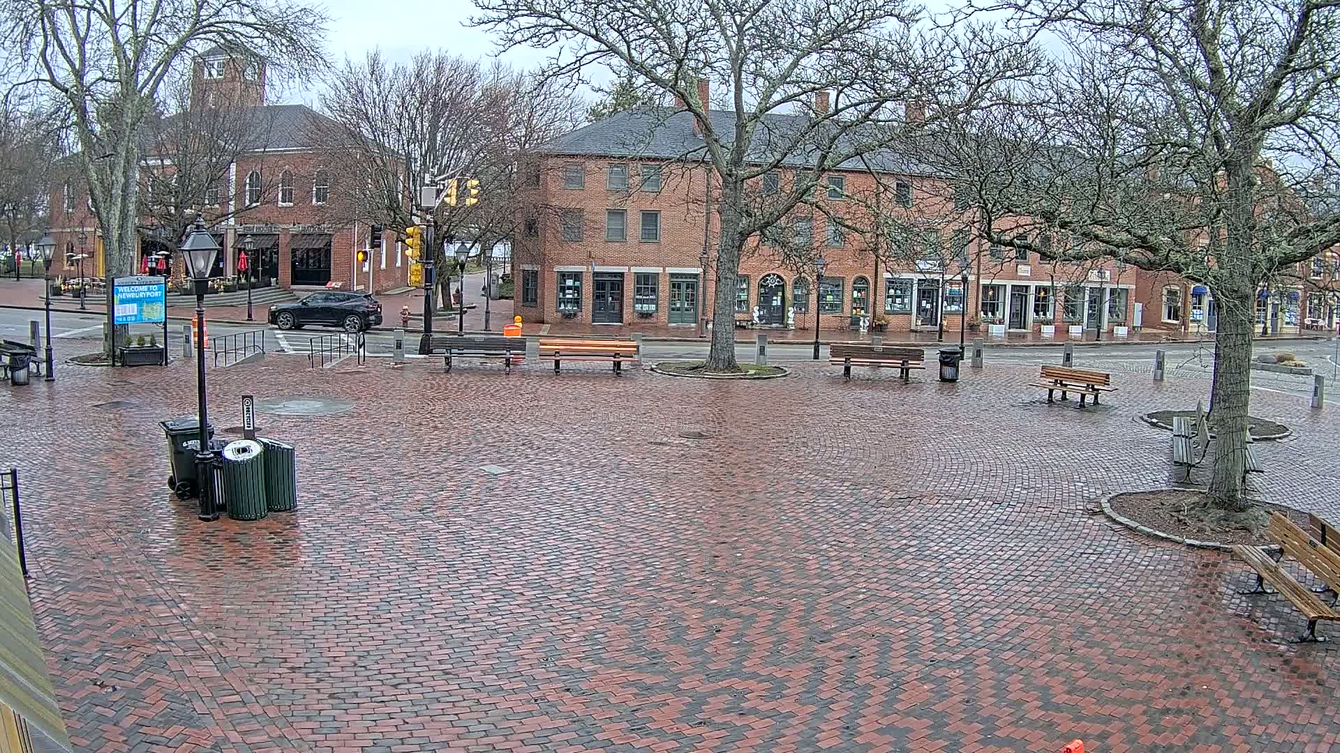 A brick town square with benches and buildings under an overcast sky, wet from recent rain.