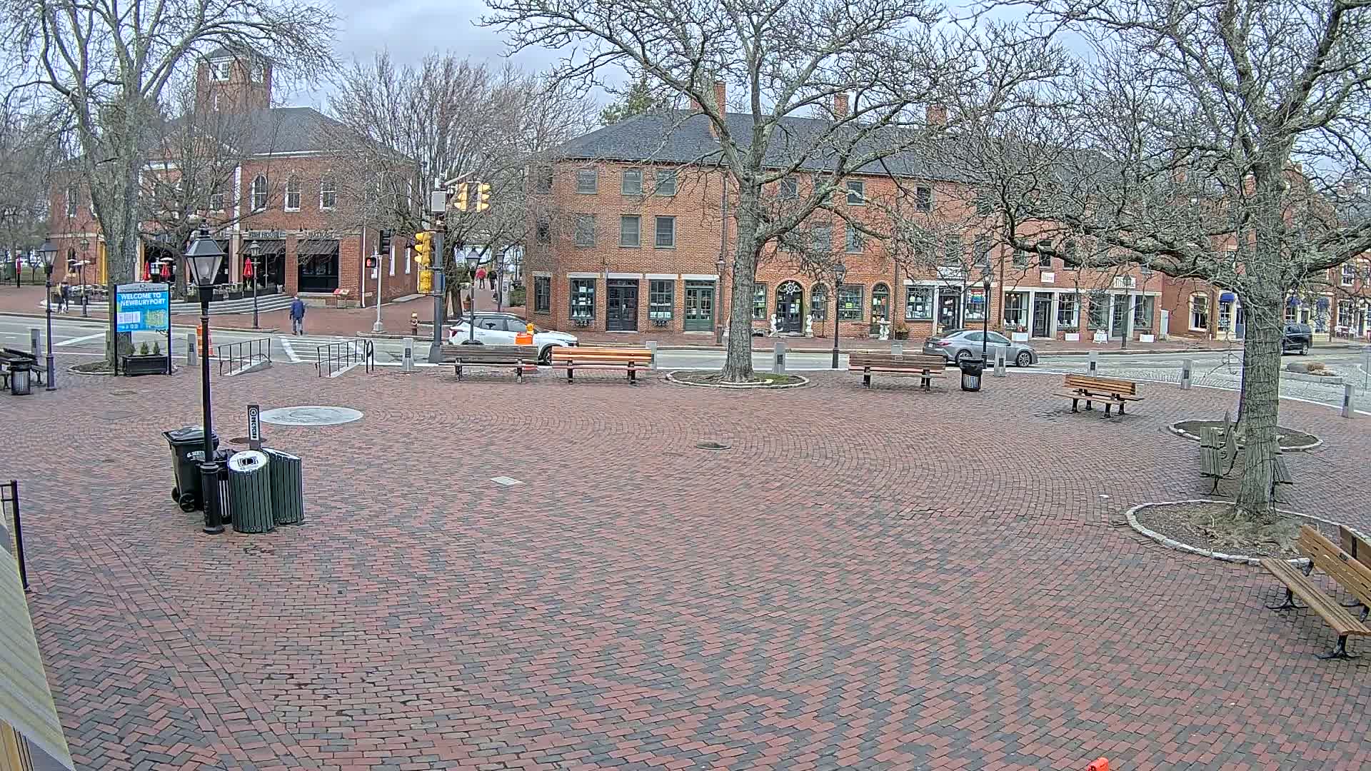 A brick plaza with several benches and a few cars is surrounded by brick buildings under a cloudy sky.