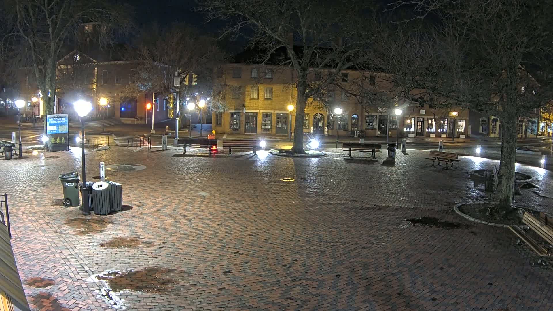 A brick town square at night, illuminated by streetlights, with a few benches and trash cans, wet from recent rain, and bare trees surrounding it.