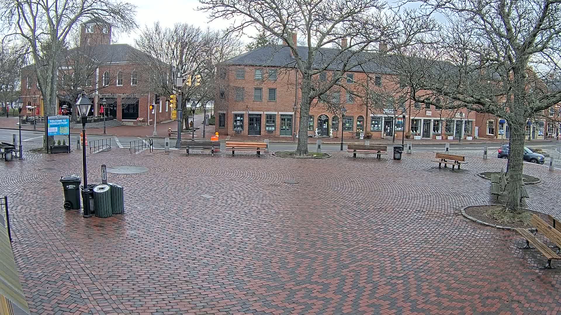 A brick town square with benches and buildings, under a cloudy sky with bare trees.