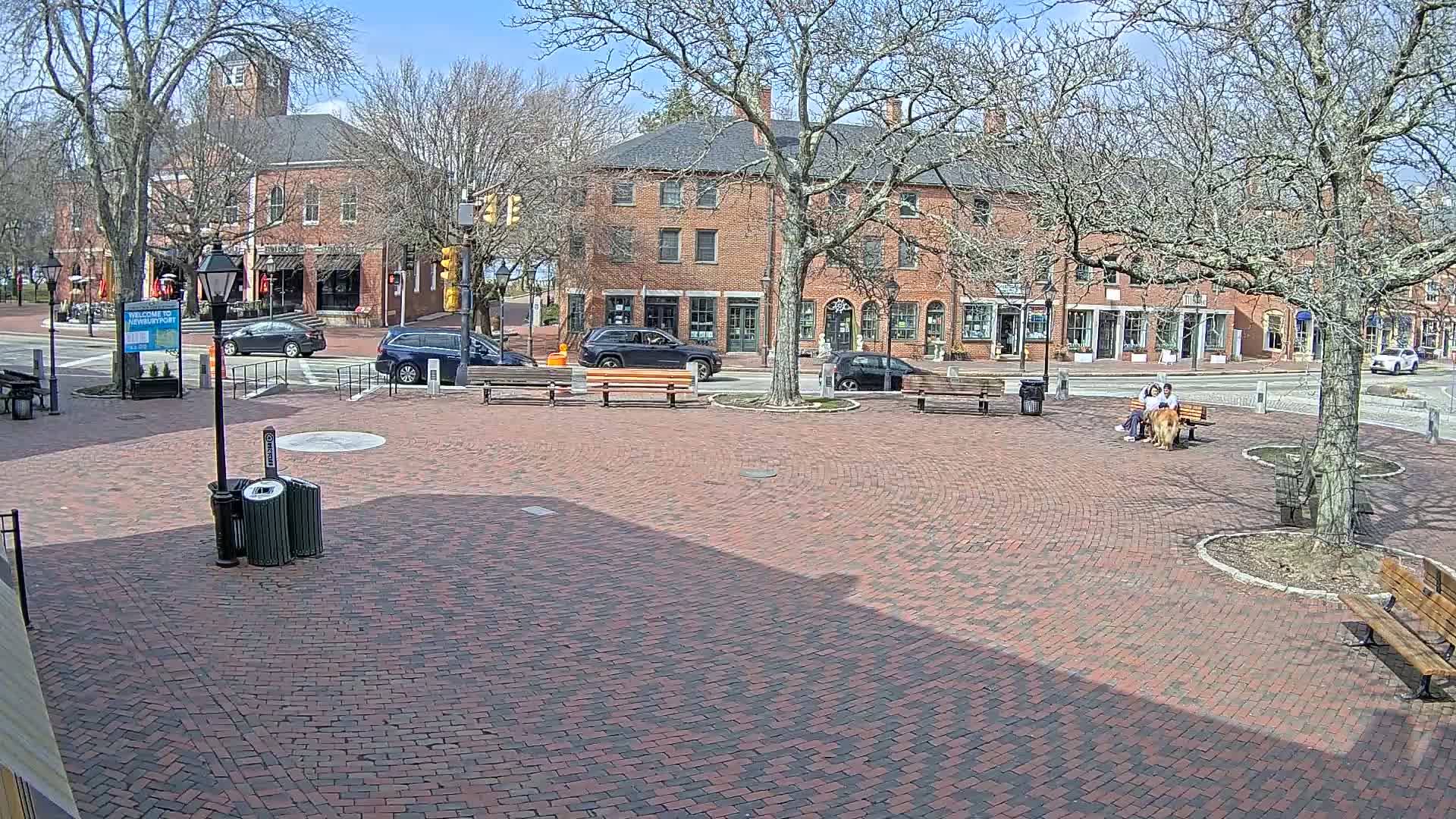 A brick town square with several cars parked nearby, two people sitting on a bench with a dog, and bare trees under a sunny sky.