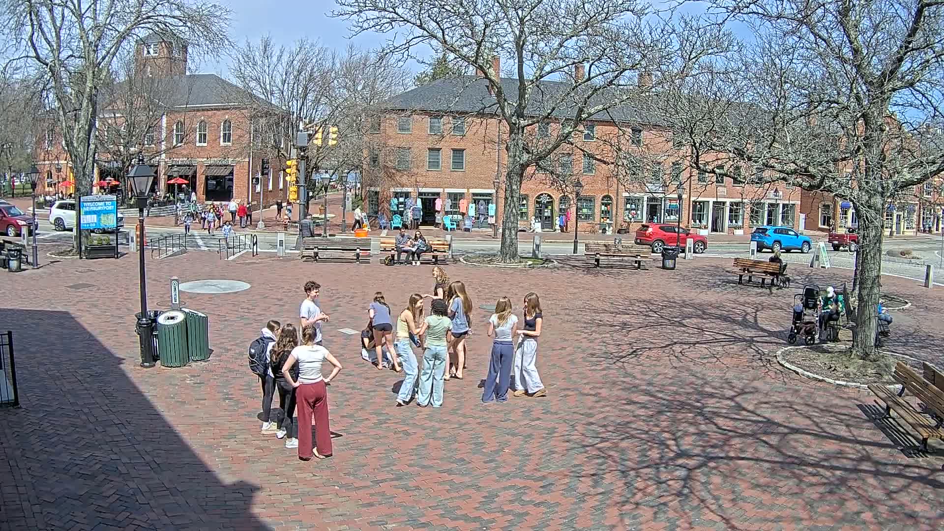 A brick town square, under a sunny sky, features several groups of young people standing and talking, while others sit on benches near brick buildings.
