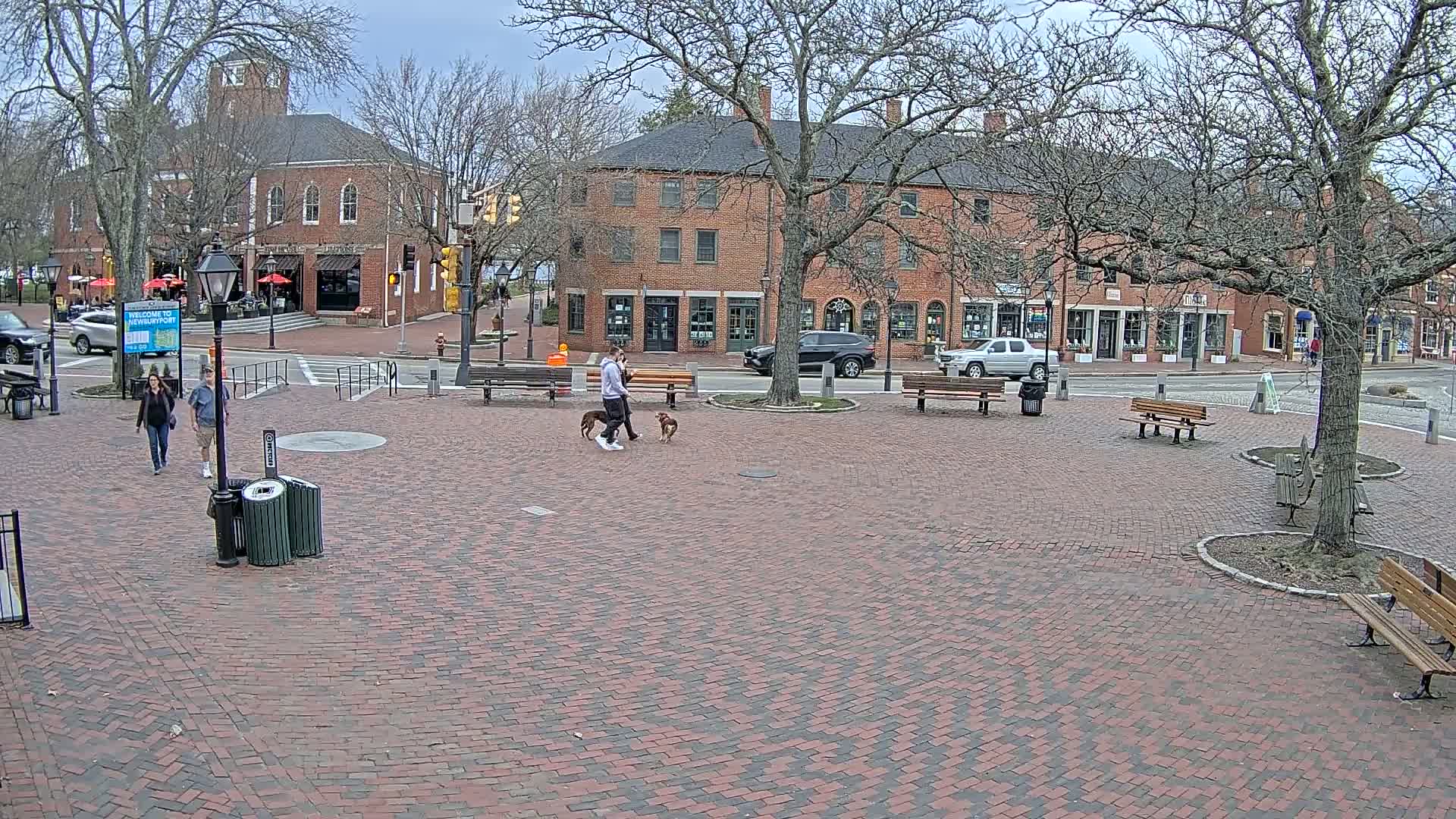 A brick plaza with benches and trees, under a cloudy sky, shows two people walking two dogs in front of brick buildings.