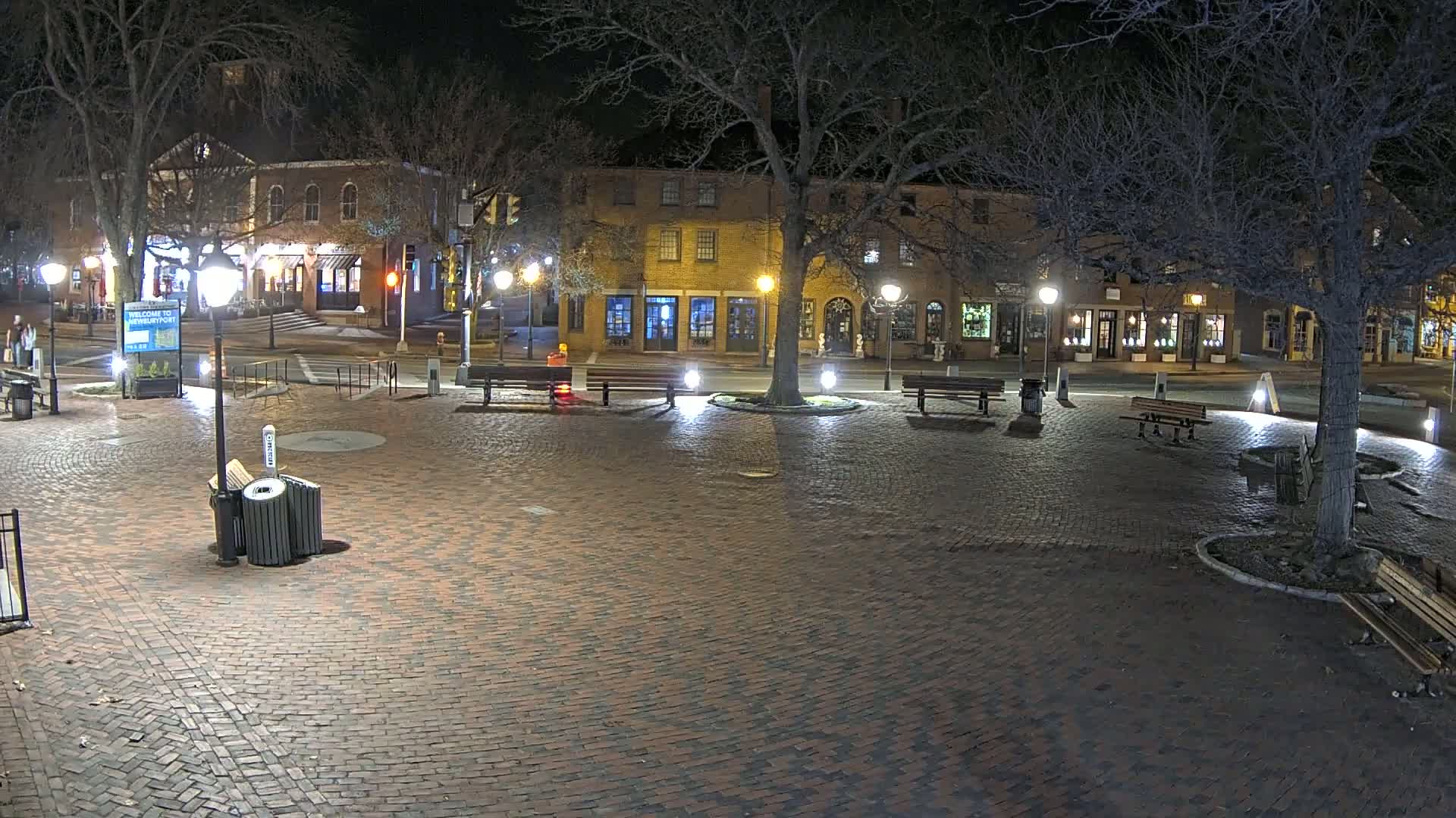 A brick town square at night, illuminated by streetlights, with several benches and trees, and buildings lining the edges.