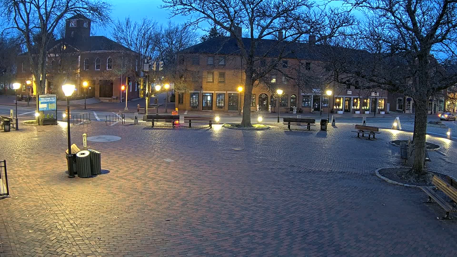 A brick town square at twilight, illuminated by streetlights, is surrounded by benches and brick buildings.