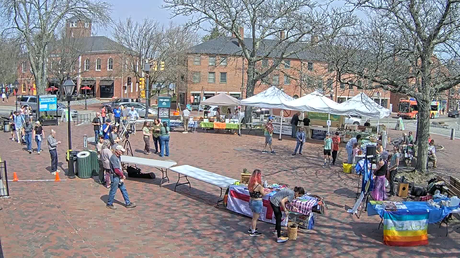 A brick paved town square hosts an outdoor market under a sunny sky, with several tents displaying merchandise and numerous people milling about.