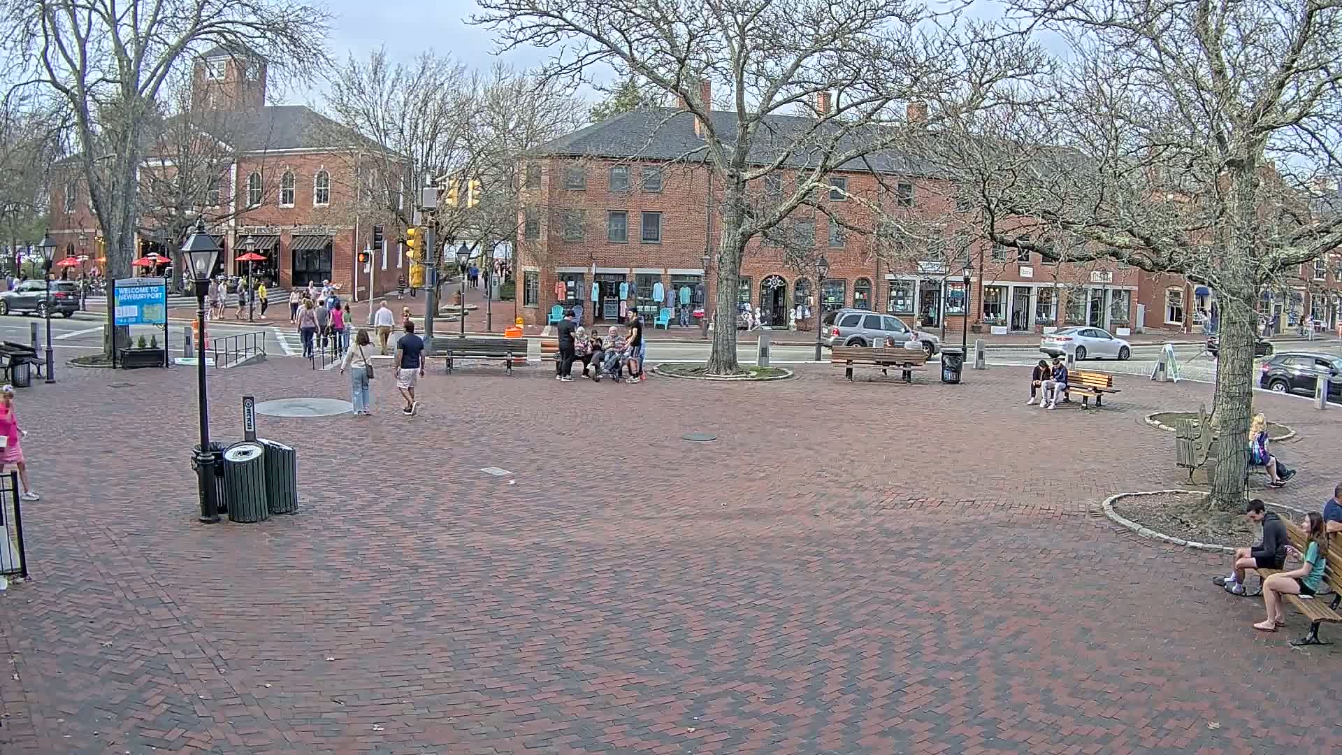 A brick town square with several people walking and sitting on benches, under a mostly cloudy sky, with brick buildings and trees surrounding the square.