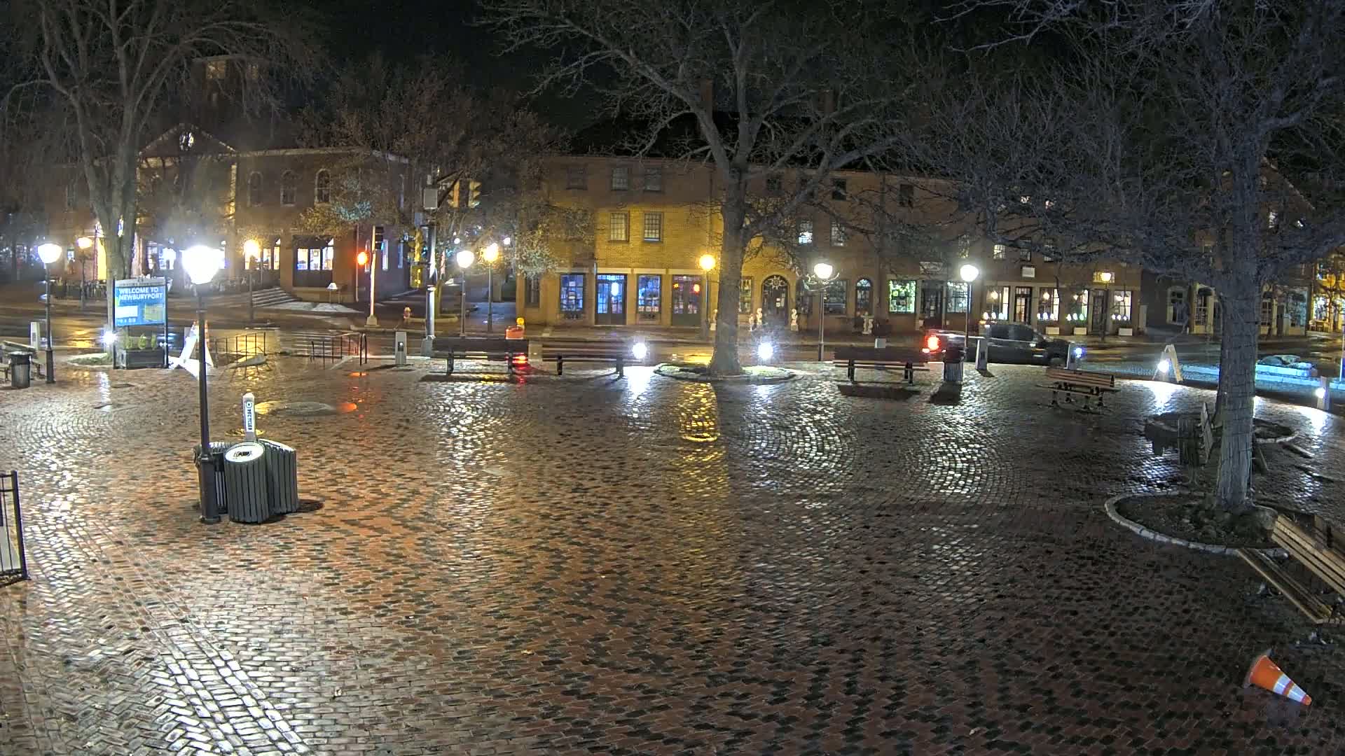 A wet, brick-paved town square at night, illuminated by streetlights, with several benches and a few cars parked nearby, surrounded by low-rise buildings.