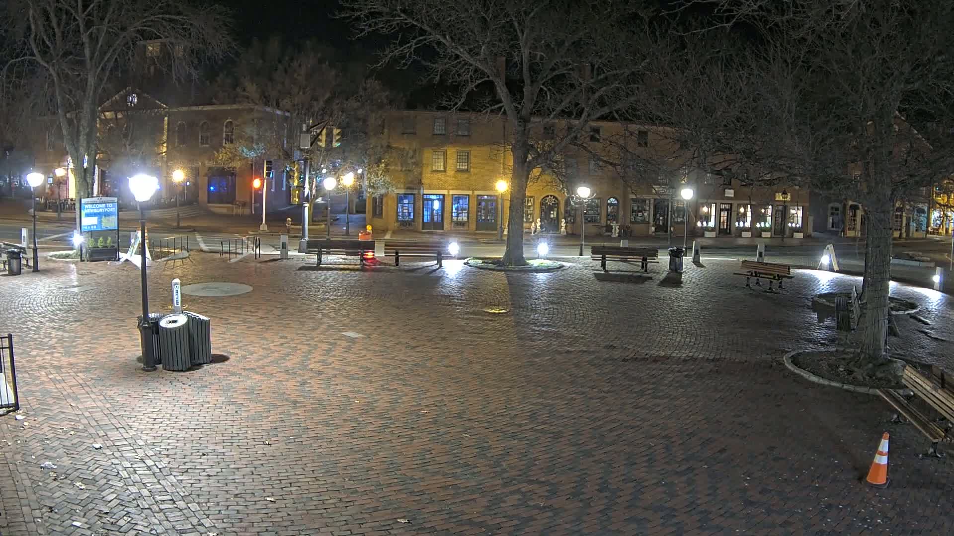 A brick town square at night, illuminated by streetlights, with benches, trees, and buildings in the background.