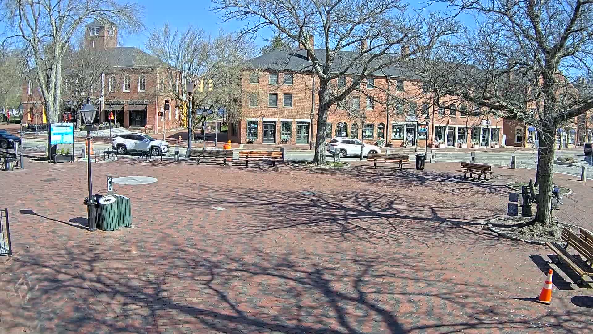 A brick town square with benches and trees, under a sunny sky, is surrounded by brick buildings.