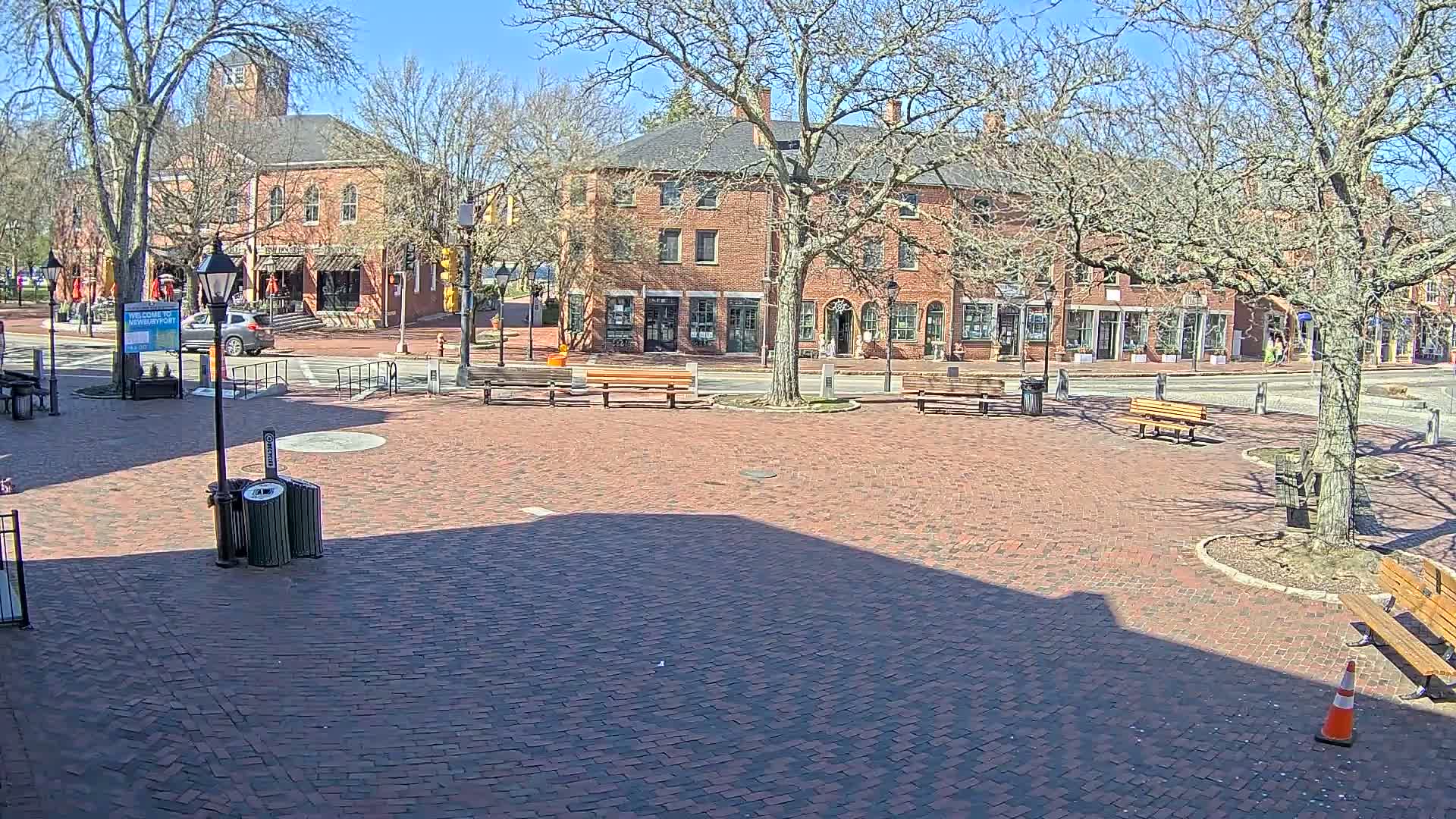 A brick town square with several benches and trees, under a sunny sky, is situated in front of brick buildings.