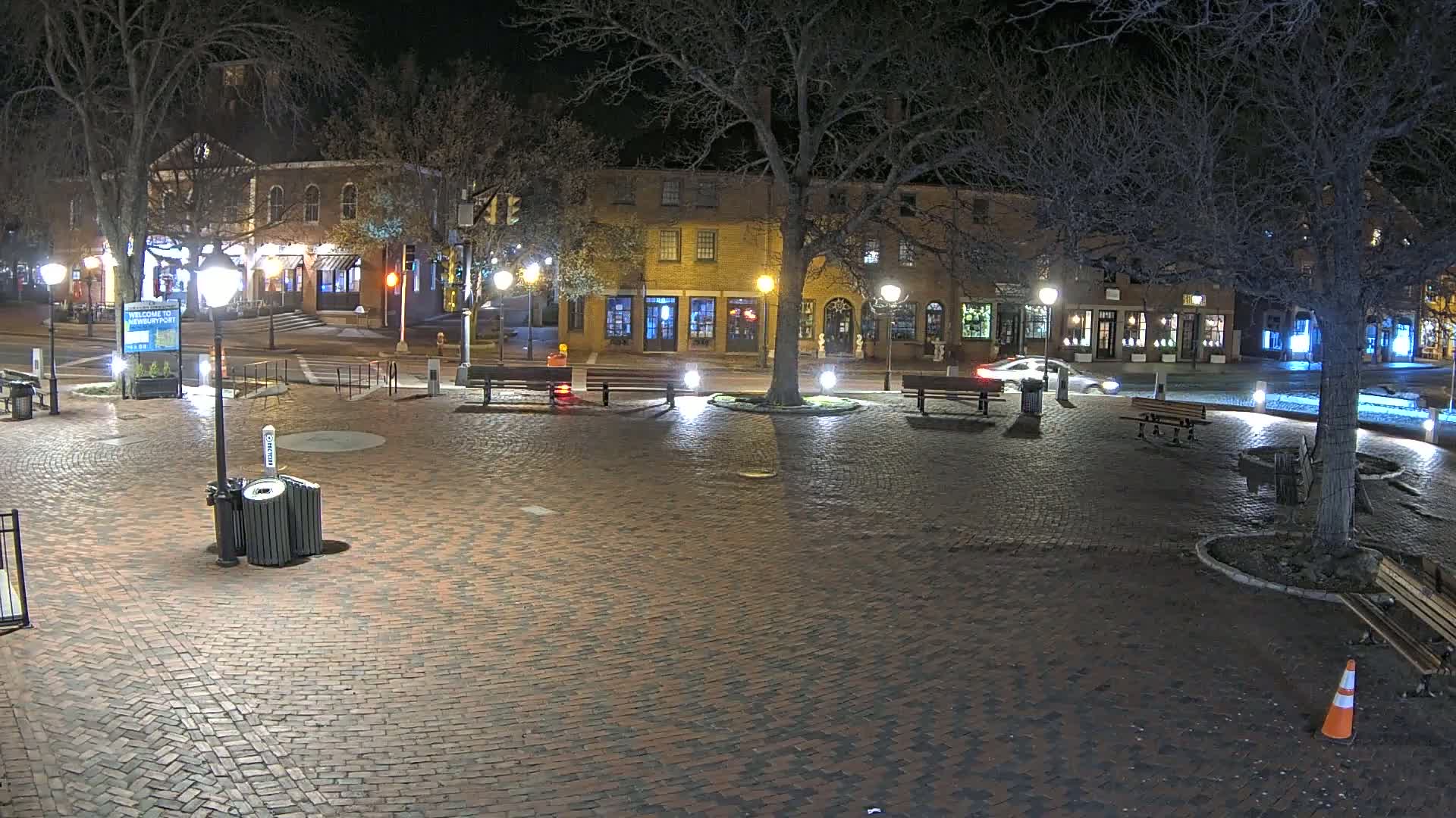 A brick plaza at night, illuminated by streetlights, is surrounded by buildings and trees with a few cars parked nearby.
