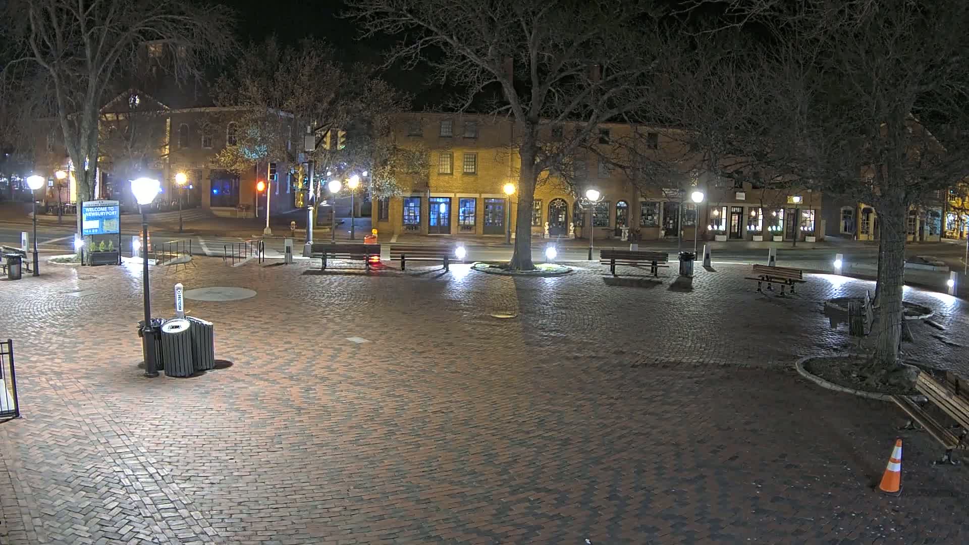 A brick town square at night, illuminated by streetlights, features benches, trees, and buildings lining the perimeter.