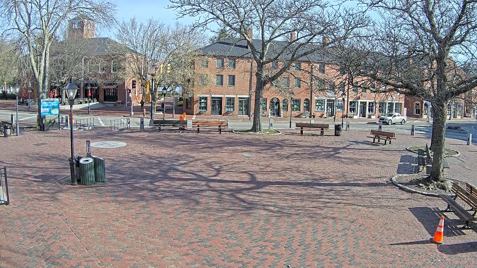 A brick plaza with benches and trees under a sunny sky sits in front of brick buildings, one of which has a clock tower.