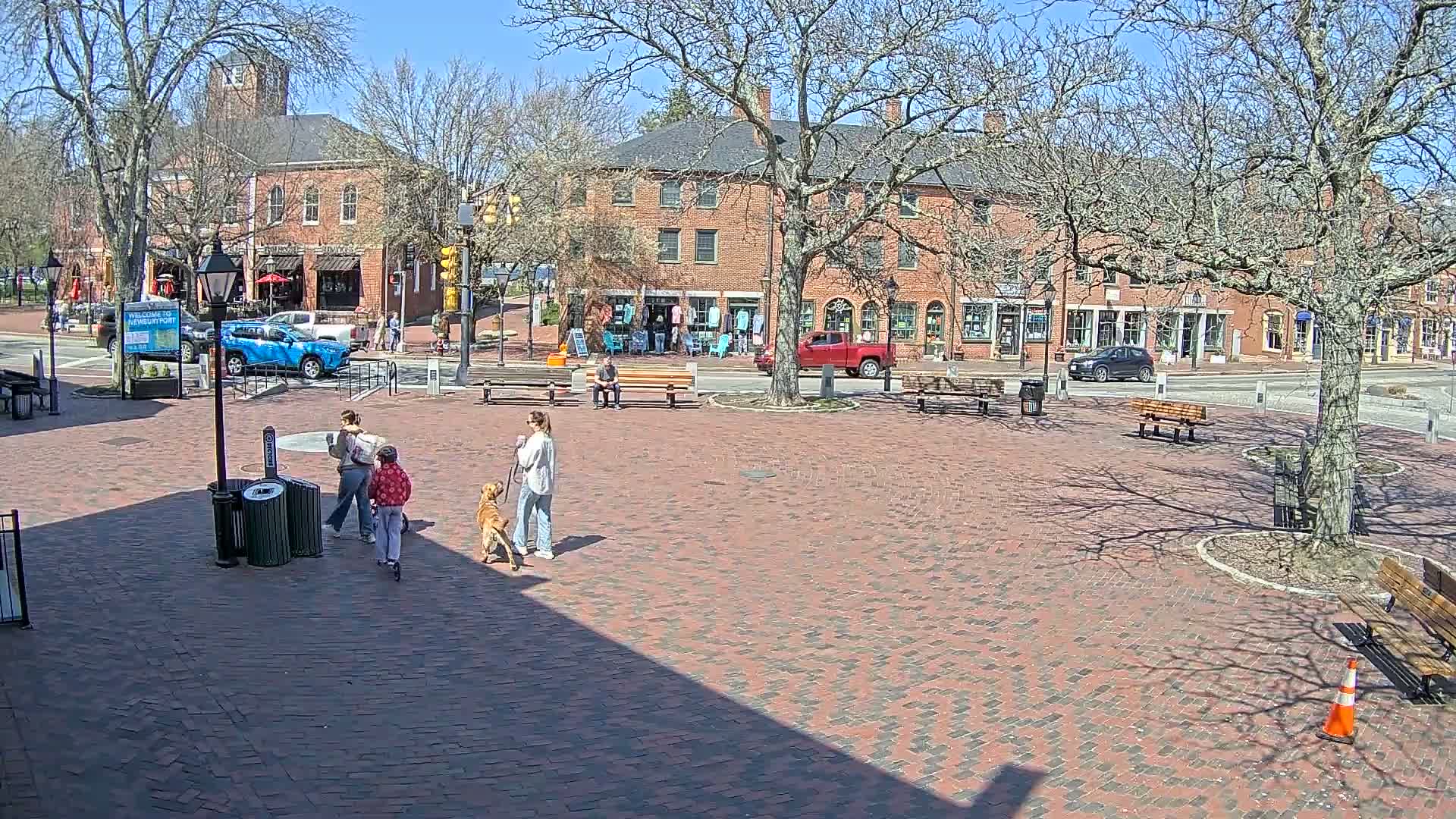 A brick town square with several people and a dog, situated in front of brick buildings under a sunny sky.