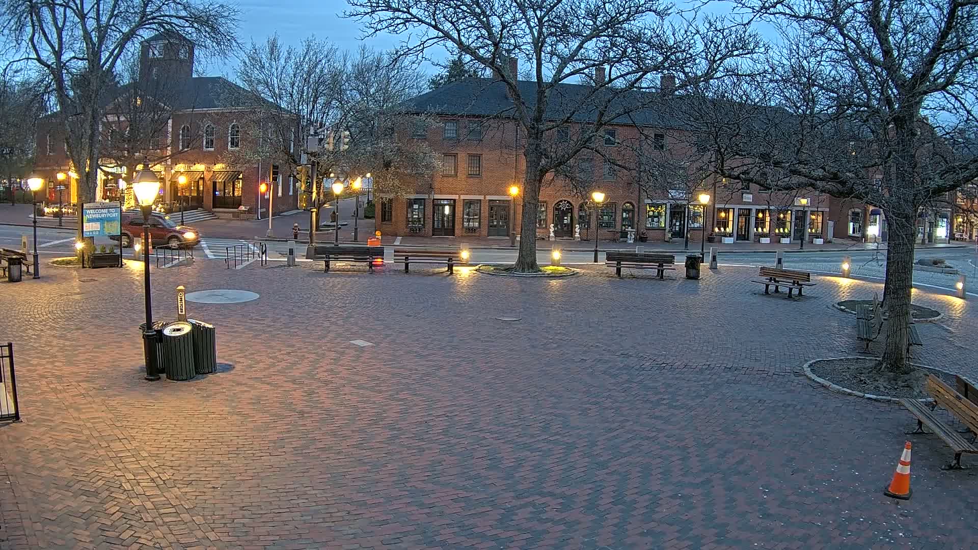 A brick town square at twilight is illuminated by streetlights, with several benches and a lone vehicle visible in the background.