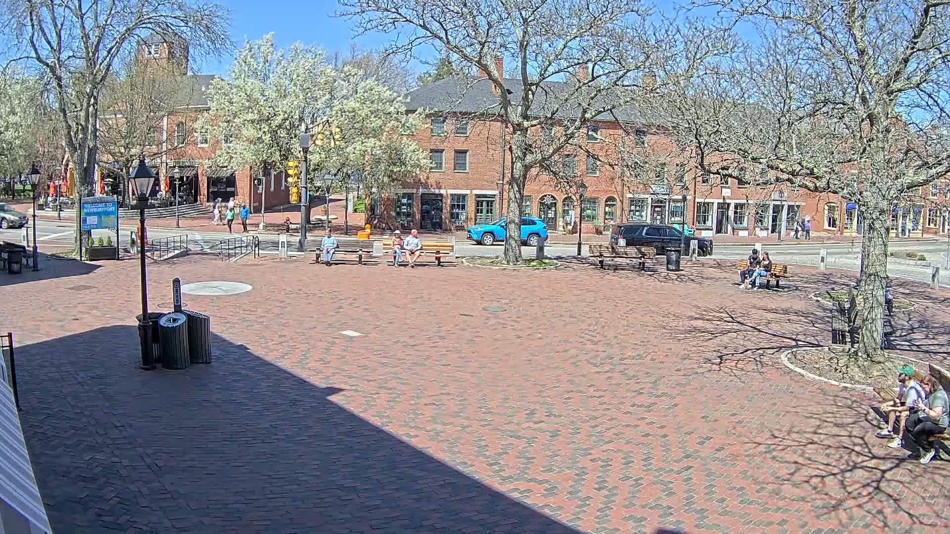 A brick town square with several people sitting on benches under sunny, clear skies, surrounded by brick buildings and leafless trees.