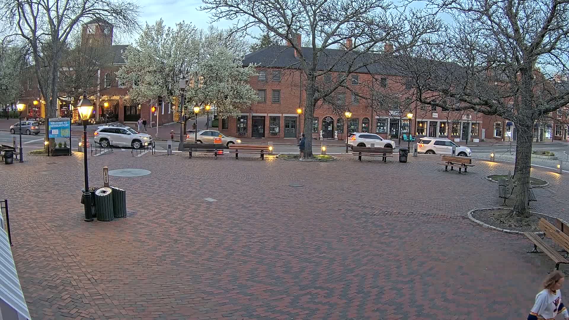 A brick plaza in the evening, with several cars parked nearby, is surrounded by brick buildings and trees in bloom under a twilight sky.