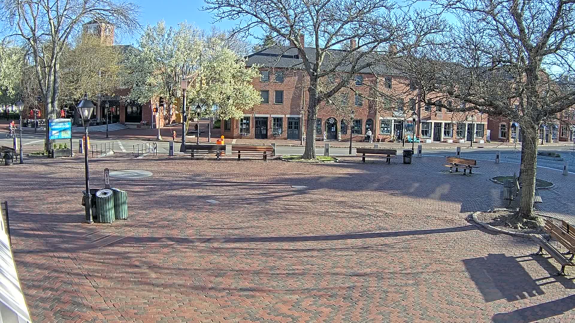 A brick town square with benches and trees under a sunny sky is situated in front of brick buildings.